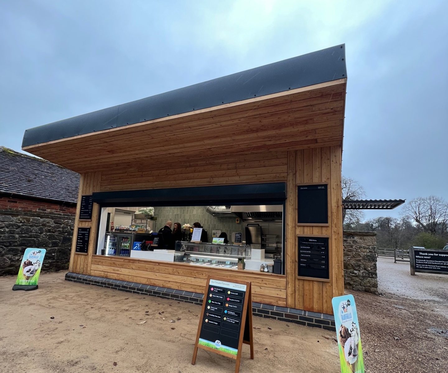 The completed BBQ kiosk at Calke Abbey: timber-clad servery with a mono-pitch roof and menu boards