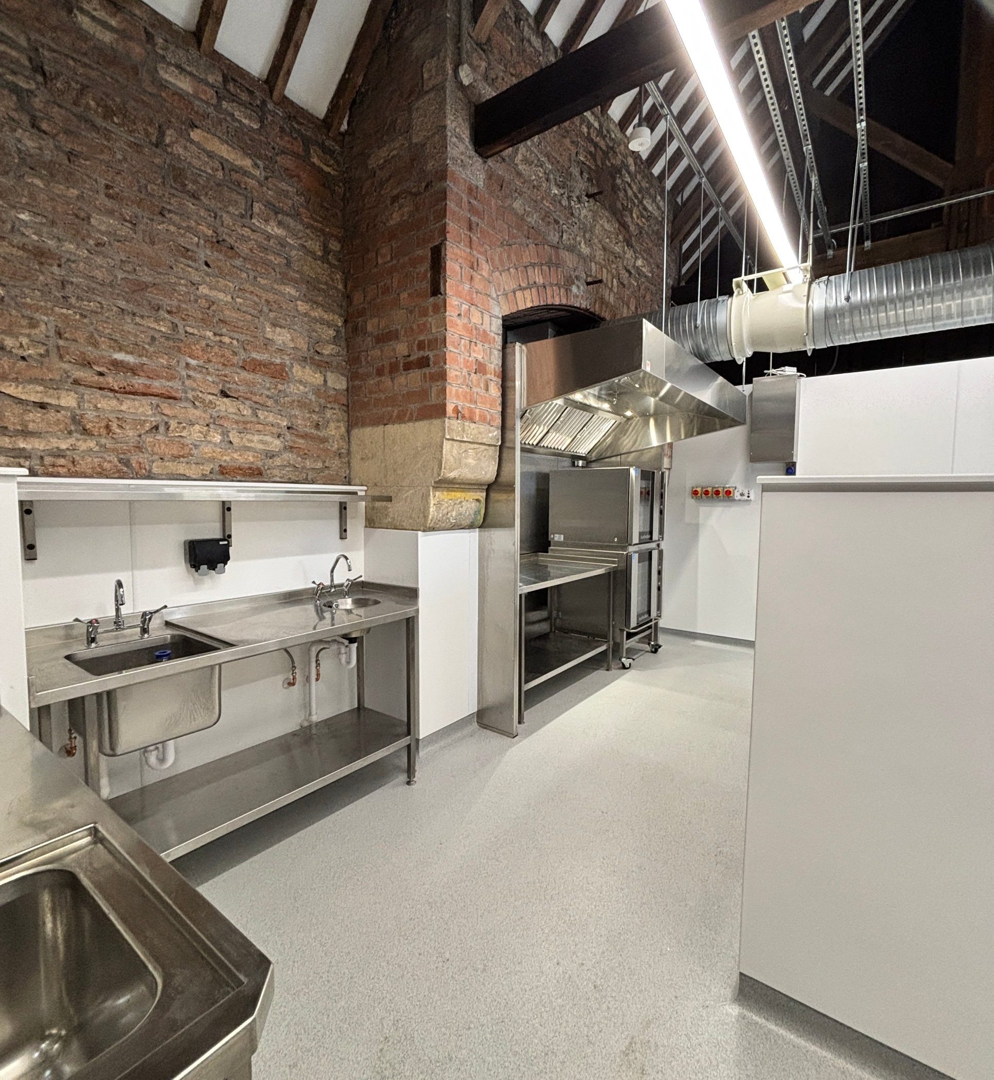 The refurbished kitchen and bakery inside the Cow Barn: new ventilation, extract canopy and servery set against the original stone and brick walls and timber roof
